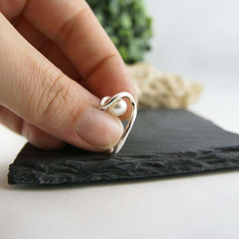 May include: A silver ring with a single white pearl. The ring is shaped like a wave and is shown on a finger.