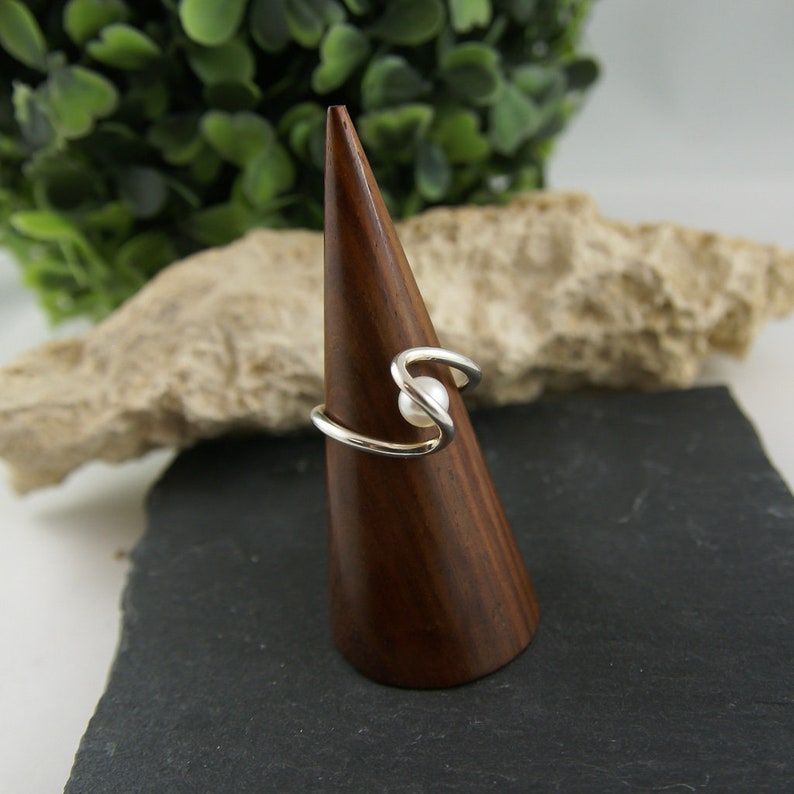 May include: A silver ring with a white pearl set in a swirl design. The ring is displayed on a wooden ring holder.