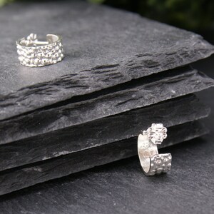 May include: Two silver ear cuffs with a textured design. The ear cuffs are on a black slate surface.