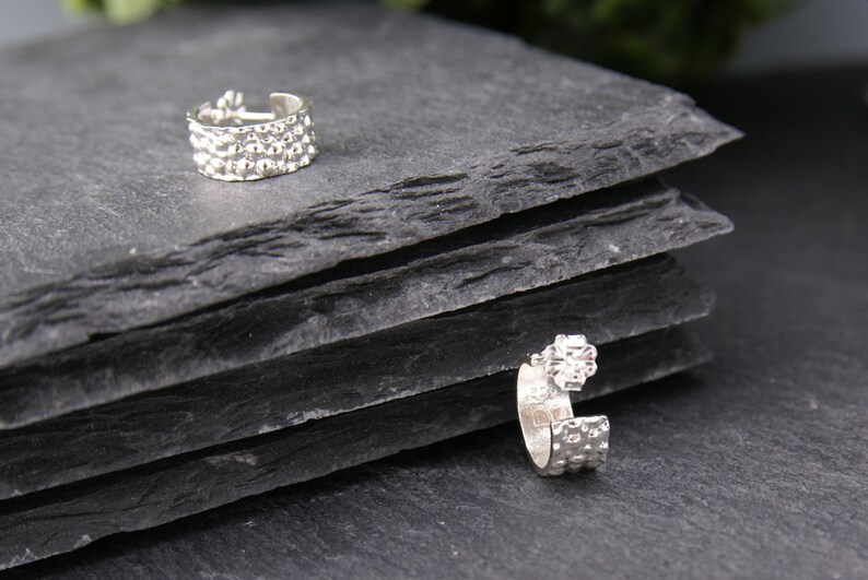 May include: Two silver ear cuffs with a textured design. The ear cuffs are on a black slate surface.