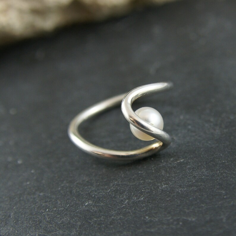 May include: A silver ring with a single white pearl set in the center. The ring is made of two bands that are intertwined.