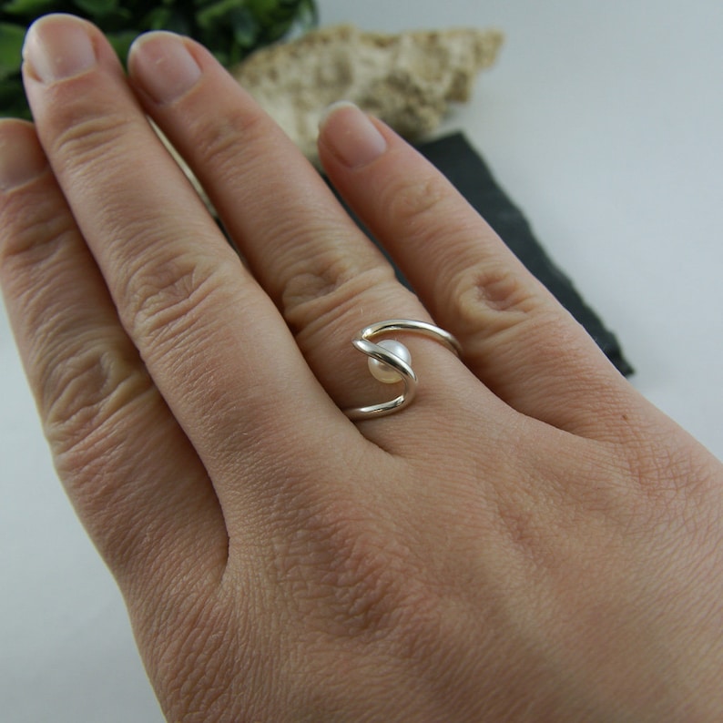 May include: A silver ring with a single pearl set in the center. The ring is designed with a curved band that wraps around the finger.