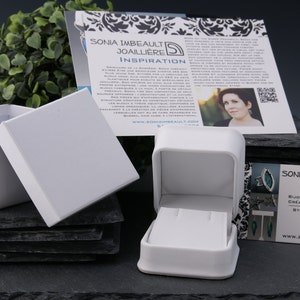 May include: A white jewelry box with a lid open, revealing a small white box inside. The box is sitting on a stack of black slate tiles. A business card with the text "SONIA IMBEAULT JOAILLIERE" is visible in the foreground.