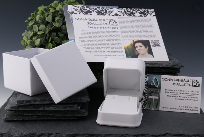 May include: A white jewelry box with a lid open, revealing a small white box inside. The box is sitting on a stack of black slate tiles. A business card with the text "SONIA IMBEAULT JOAILLIERE" is visible in the foreground.