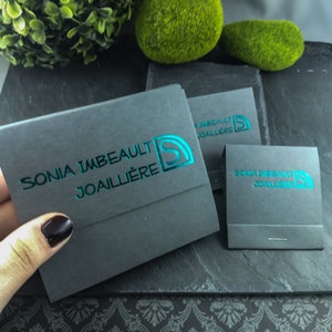 May include: Two gray boxes with the text "SONIA IMBEAULT JOAILLI&Egrave;RE" in teal lettering. The boxes are on a black surface with a decorative pattern.