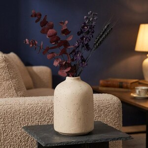 May include: A cream-colored ceramic vase with a speckled finish containing a bouquet of dried dark purple flowers, deep red eucalyptus leaves, and dark wheat stalks, creating a contrast between the light vessel and the dark foliage.
