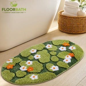 May include: An oval-shaped bath mat featuring a textured green surface with a pattern of white and orange flowers and green lily pads. The rug has a soft, tufted pile construction and a finished edge, designed for placement on a bathroom floor.