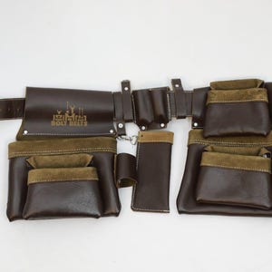 May include: A dark brown leather tool belt featuring multiple pouches with olive green suede accents. The belt includes a metal buckle, several pockets of varying sizes, and a logo stamped on the left pouch that reads BOLT BELTS in white.