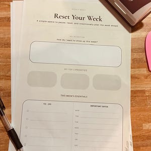 May include: A white weekly planner sheet titled Reset Your Week with sections for intentions, top three priorities, to-do lists, and important dates. A black ballpoint pen, a maroon and white hole punch, and a pink heart-shaped sticky note are included.