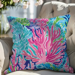May include: A square decorative pillow with a vibrant coral reef design. The pillow features a colorful pattern of pink, blue, and green coral against a dark blue background. The pillow is placed on a cream-colored tufted sofa.
