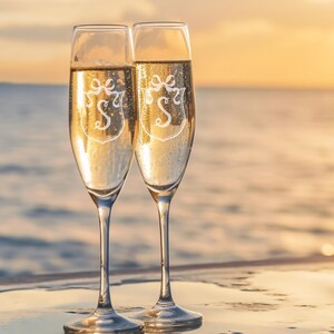 Puede incluir: Dos copas de champán llenas de líquido burbujeante, cada una grabada con un monograma blanco y un diseño de lazo. Las copas se colocan sobre un fondo de puesta de sol sobre el mar, creando un ambiente festivo.