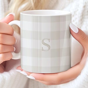 May include: A white ceramic mug with a light gray plaid pattern and a large letter "S" in the center. The mug has a white handle and is held by a person wearing a white sweater.