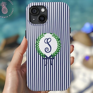 May include: A navy and white striped phone case featuring a green wreath with a blue bow and the letter "S". The phone case has a camera cutout and is held in a hand. The Summer Market logo is in the upper left corner.