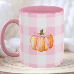 Puede incluir: Una taza de cerámica con asa e interior rosas. El exterior presenta un estampado de cuadros vichy rosa y blanco y una ilustración de acuarela de una calabaza naranja con un lazo rosa. La taza está sobre una superficie de madera.