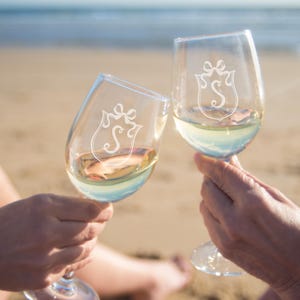 May include: Two clear wine glasses filled with white wine are held up in a toast. Each glass has a decorative crest with the letter "S" etched on the front. The background is a sandy beach with the ocean in the distance.