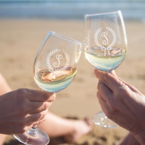 May include: Two clear wine glasses filled with white wine are held up in a toast. Each glass has a decorative wreath design with the letter "S" etched on the glass. The background is a sandy beach with the ocean in the distance.
