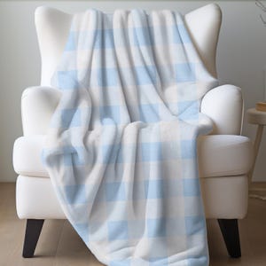 May include: A soft, plush blanket in a light blue and white checkered pattern is draped over a white armchair. The blanket's texture appears cozy and inviting, perfect for adding warmth and style to a living space.