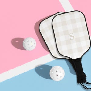 Op de afbeelding: Twee pickleball rackets met een beige en wit geblokt patroon en zwarte rand, samen met twee witte geperforeerde pickleballs. De rackets hebben een zwart handvat en de letter "S" op het racketblad. De achtergrond is roze en blauw.