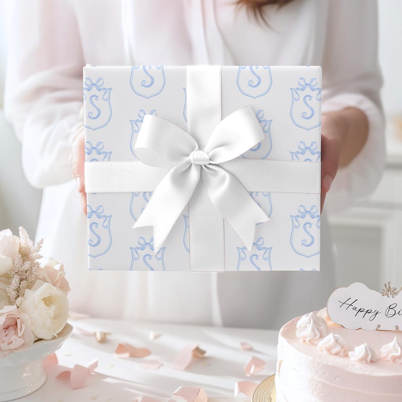 May include: A wrapped gift box with a white ribbon and bow. The wrapping paper is white with a light blue pattern of a crest with the letter "S". The gift is presented on a white surface, with a cake and flowers in the background.