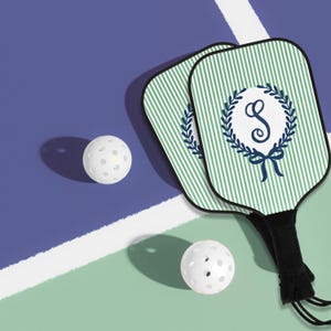 Könnte beinhalten: Zwei Pickleball-Schläger mit grün-weißen Streifen und einem marineblauen Monogramm "S" in einem Kranz. Zwei weiße Pickleballs sind ebenfalls auf einem blau-grünen Platz zu sehen.