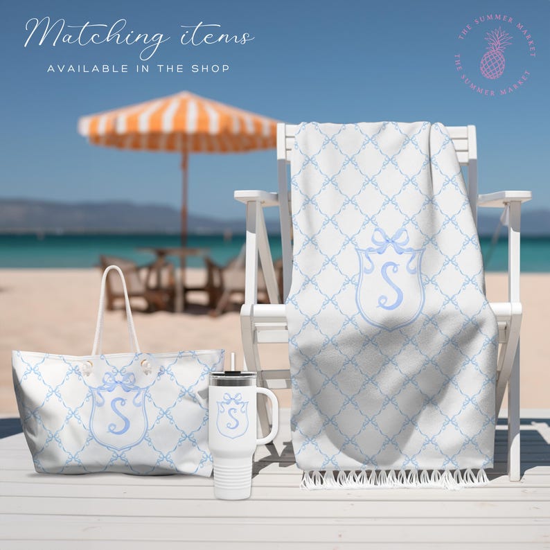 May include: White beach tote bag, towel, and travel mug with a light blue bow and initial "S" design. The items are on a white wooden surface with a beach umbrella and ocean in the background. Text on the image says "Matching items available in the shop."