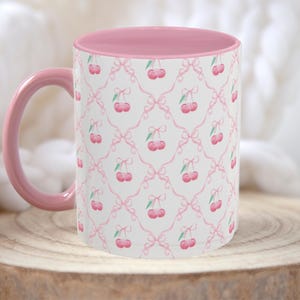 May include: Pink ceramic mug with a white background and a repeating pattern of pink cherries and bows. The mug has a pink interior.