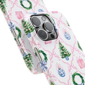 May include: White phone case with a repeating pattern of pink bows, Christmas trees, gift boxes, wreaths, and blue and white ornaments. The case is designed to fit a smartphone and has cutouts for the camera and buttons.