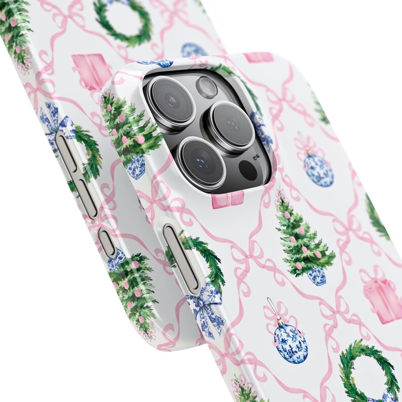 May include: White phone case with a repeating pattern of pink bows, Christmas trees, gift boxes, wreaths, and blue and white ornaments. The case is designed to fit a smartphone and has cutouts for the camera and buttons.