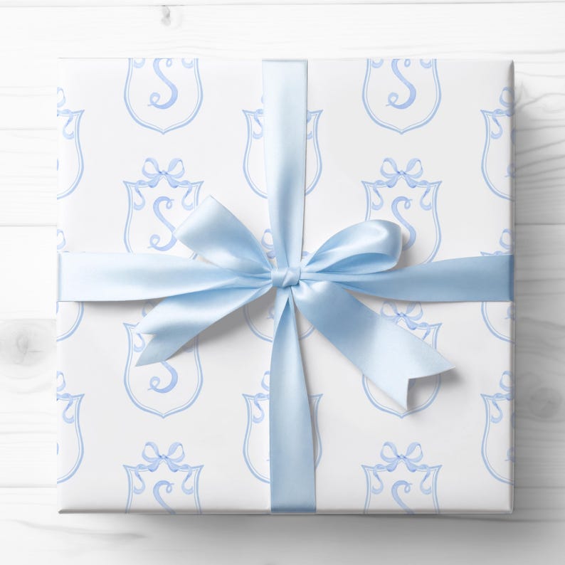 May include: A wrapped gift with white paper featuring a repeating light blue crest design and a matching satin ribbon tied in a bow. The gift is ready to be given.