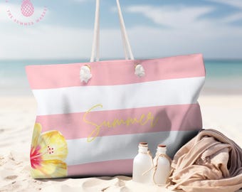 May include: Pink and white striped tote bag with rope handles. The bag features a yellow hibiscus flower and the word "Summer" in yellow script. Two small white bottles and a tan towel are on the sand.