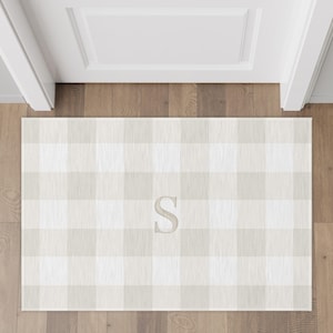 May include: A rectangular doormat with a large, light beige and white buffalo check pattern. The letter "S" is embroidered in the center of the mat. The mat is placed in front of a white door on a hardwood floor.