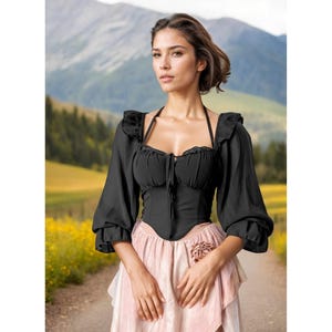 May include: A black corset-style top featuring long, puffed sleeves with ruffled shoulders and cuffs, a sweetheart neckline with a central tie detail, and a structured bodice paired with a layered, light pink skirt adorned with a fabric flower.