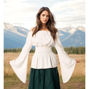 May include: An off-the-shoulder white blouse featuring long, wide bell sleeves and a smocked, elasticized waistline that creates a gathered, peplum-style hem. The garment is paired with a solid dark green skirt that reaches below the waist.