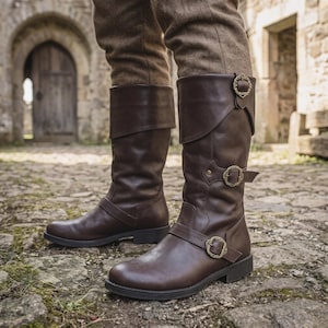 May include: A pair of dark brown leather mid-calf boots featuring a folded cuff design, a rounded toe, and two adjustable straps across the shaft and ankle. Each strap is secured with an ornate, antique-style brass buckle with intricate detailing.