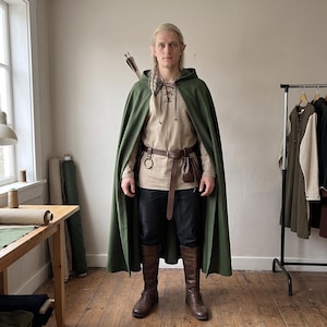 May include: A hooded olive green cloak worn over a beige long-sleeved tunic with a lace-up neckline, black trousers, and brown leather boots. A brown leather belt with a small pouch and a metal ring is worn at the waist, with an arrow quiver visible.