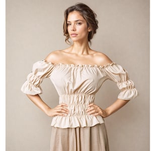 May include: A beige off-the-shoulder blouse featuring a gathered elastic neckline, puffed elbow-length sleeves with elasticized cuffs, and a wide smocked waist panel that creates a ruffled hemline. The garment is made of a solid light-colored fabric.