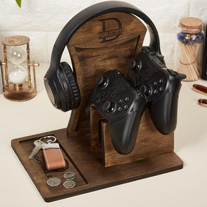 Könnte beinhalten: Ein dunkelbrauner Gaming-Organizer-Ständer aus Holz mit einer vertikalen Stütze, die mit dem Buchstaben D und dem Namen DONALD graviert ist. Der Ständer hält schwarze Over-Ear-Kopfhörer und zwei schwarze Gamecontroller sowie eine Ablage.