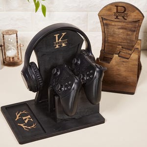 Könnte beinhalten: Ein schwarzer Gaming-Organizer-Ständer aus Holz mit Kopfhörerhalterung, Controller-Halterung und einer Basisablage mit eingraviertem Buchstaben K und Geweihmotiv. Der Ständer zeigt den Namen KERWIN. Ein zweiter brauner Holzständer trägt den Namen ROBERT.