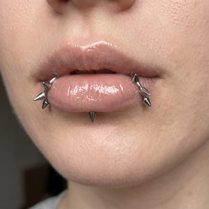 Pode incluir: Um conjunto de joias para piercing labial em tom prateado, apresentando uma barra curva com várias pontas cónicas afiadas. A joia é posicionada ao redor do lábio inferior, com dois segmentos curvos nas laterais e uma ponta central na base.