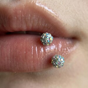 Può includere: Un piercing al labbro con due borchie sferiche, ciascuna incastonata con molteplici strass iridescenti e multicolori che catturano la luce. Il gioiello è composto da una sfera superiore e una inferiore, entrambe con una superficie riflettente.