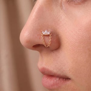 May include: A gold-toned nose ring featuring a decorative top element with three marquise-cut clear stones arranged in a fan shape, connected by a delicate metal chain that loops downward to create a draped effect on the side of the nostril.