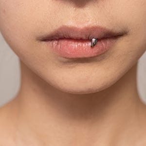 May include: A silver-colored, curved, fang-shaped lip piercing jewelry piece positioned on the lower lip. The metal accessory has a smooth, polished finish and a pointed end that rests against the skin, creating a distinct, singular visual accent.