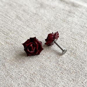 Op de afbeelding: Een paar donkerrode roosvormige oorknopjes met een metalen stift en een platte ronde achterkant. De rozenblaadjes zijn gevormd in diepe bordeauxrode tinten met een gladde afwerking, bevestigd aan een zilverkleurige metalen stift en achterkant.