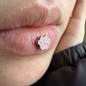 Könnte beinhalten: Ein silberfarbener Metall-Körperschmuck in Form eines Pfotenabdrucks. Die Mitte des Pfotenballens und die vier Zehen sind mit einer hellrosa, glitzernden Emaille-Einlage gefüllt. Der Schmuck befindet sich auf der Unterlippe und hat ein metallisches Finish.