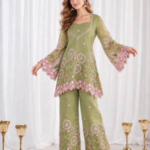 May include: An olive green tunic and wide-leg trousers set featuring intricate pink floral embroidery with scalloped edges. The tunic has long, flared sleeves and a square neckline adorned with decorative trim, while the pants feature matching floral patterns.