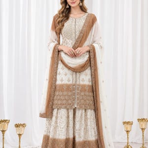 May include: A white and tan three-piece ensemble featuring a long-sleeved tunic with intricate gold embroidery, a matching flared skirt with a wide embroidered hem, and a sheer dupatta with a tan border. The Liora Couture logo is visible.