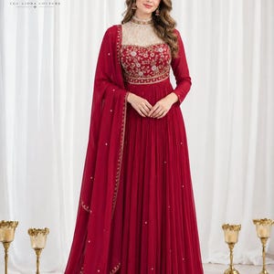 May include: A floor-length deep red gown featuring a high-neck bodice with intricate gold floral embroidery and a sheer cream-colored yoke. The skirt is pleated, and the ensemble includes a matching red dupatta with gold border detailing.