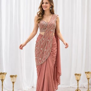 Può includere: Un sari color rosa polvere composto da una blusa senza maniche e una gonna drappeggiata. Il capo è decorato con intricate perline argentate e ricami di paillettes disposti in un motivo a reticolo vorticoso sul corpetto e sul pallu.