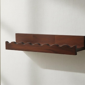 Op de afbeelding: Een wandplank van hout met een donkerbruine afwerking en een opvallende, geschulpte voorrand. De plank heeft een plat achterpaneel en een horizontale basis, ontworpen met een golvende, decoratieve rand aan de voorzijde voor een textuur.