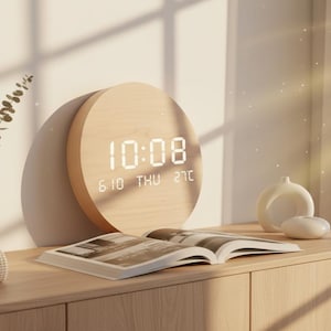 May include: A circular light-colored wood digital clock displaying the time 10:08, the date 6 10, the day THU, and the temperature 27C in white LED digits. The clock is positioned on a wooden surface next to an open book and decorative items.